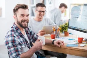 40227691 - getting the job done. handsome young man holding a cup of coffee and smiling while sitting at desk in the office with his colleagues working in the background